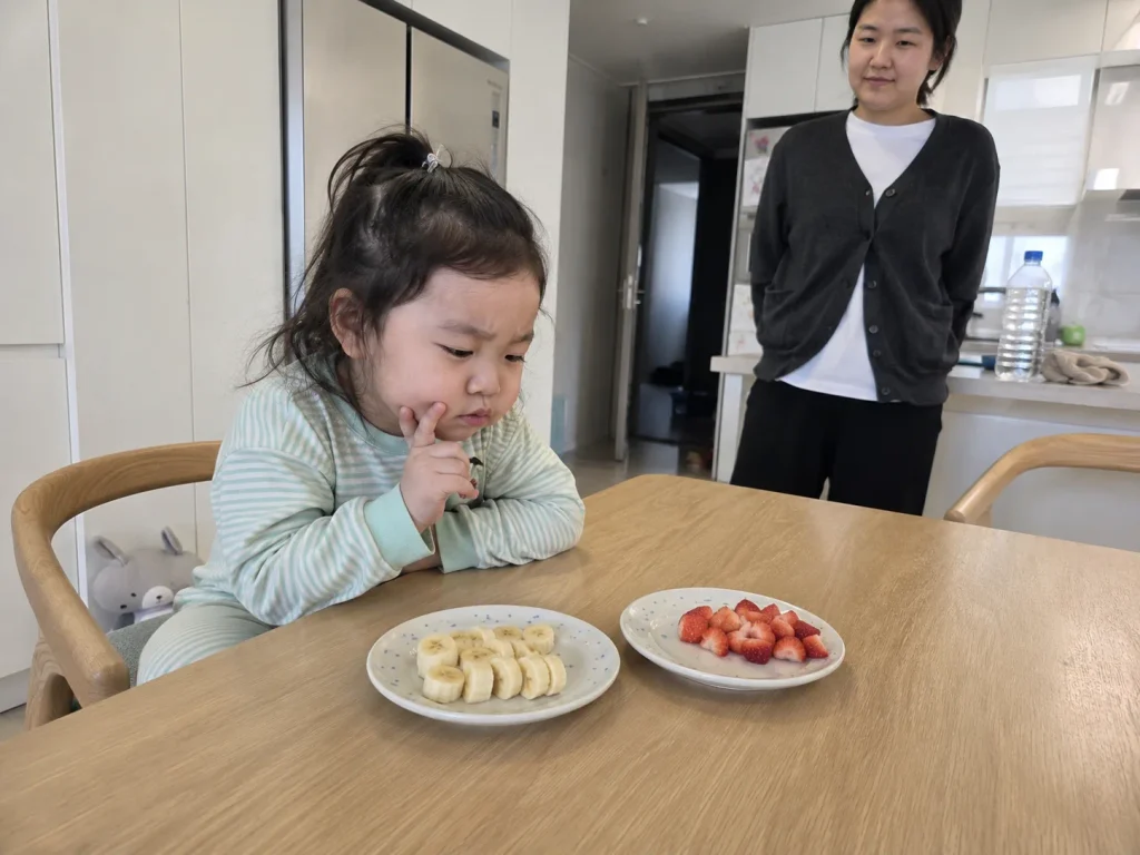 바나나와 딸기 두 접시 중 무엇을 먹을지 고민하는 아이