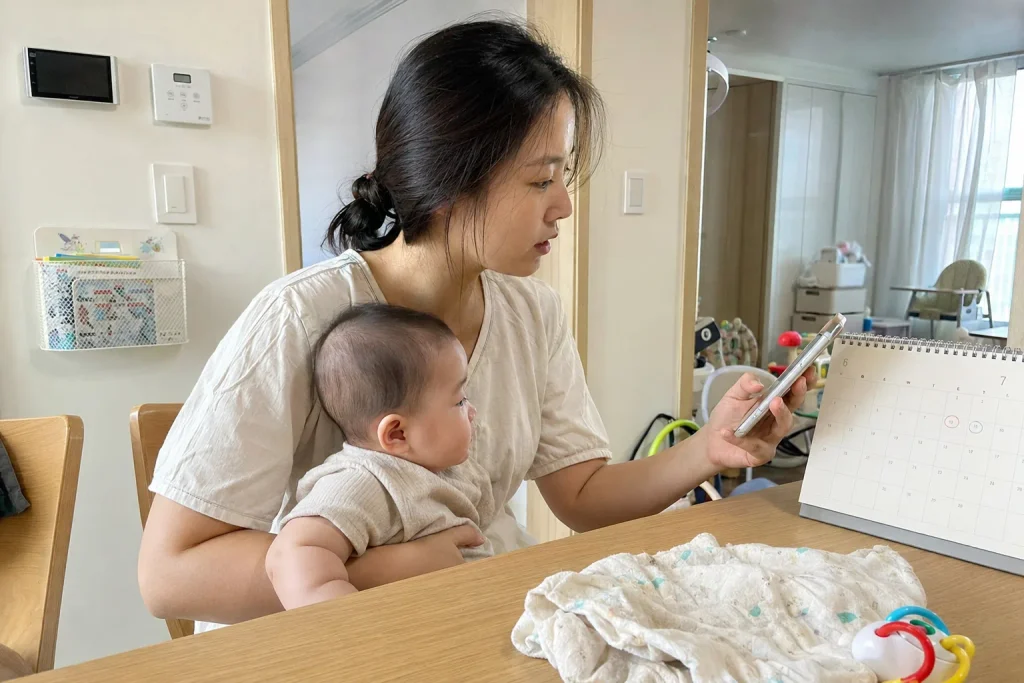 아기를 안고 휴대폰과 달력으로 출생일 기준 지원금을 확인하는 엄마