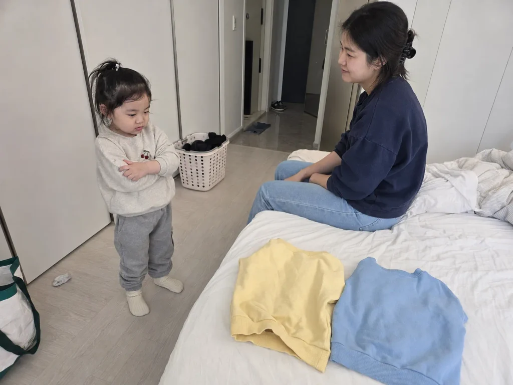 침대 위 옷 두 벌 중 무엇을 입을지 고민하는 아이와 기다려주는 엄마